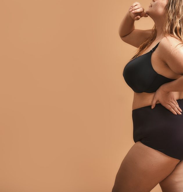 Side view of plus size sexy model in black lingerie standing against brown wall in studio